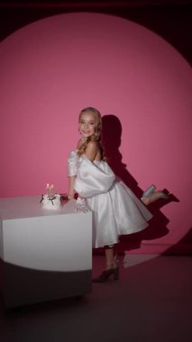 Beyaz elbiseli ve topuklu ayakkabılı gülümseyen genç kız stüdyoda doğum günü pastasıyla poz veriyor..