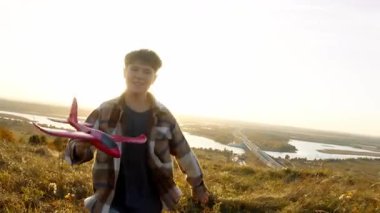 Orta boy yakın çekim, akşam güneşi sonbahar tarlasında ve arka planda ağaç dalları arasında akarken oyuncak uçağı kaldırırken, neşeli bir şekilde kameraya doğru koşan çocuğu gösteriyor..