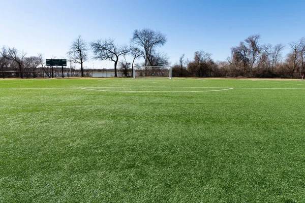Soccer goal on a field with artificial grass in a city park with trees and a lake in the background.