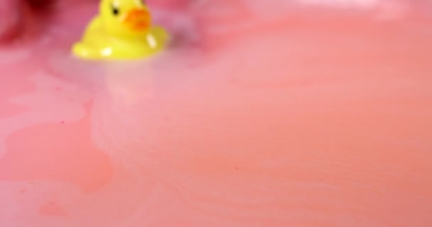 La mano femenina juega con un juguete de pato amarillo de goma en un baño caliente lleno de una espuma de color rosa. — Vídeo de stock