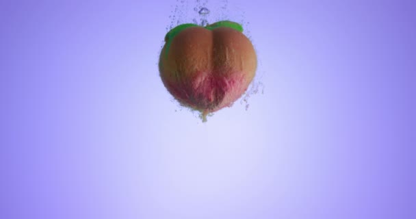 Colorida bomba de baño con una forma de melocotón cayendo en agua limpia sobre fondo púrpura claro. Filmado en cámara lenta con cámara RED — Vídeo de stock