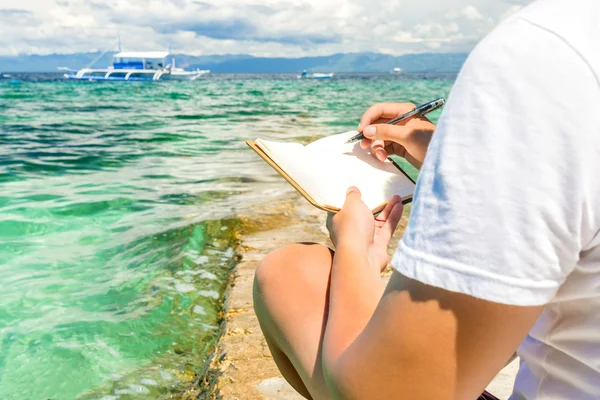 Blanke man zit op de golfbreker van tropische turquoise zee en wrigting door de pen in de notitie pad met Filippijnse boot achtergrond op zonnige zomerdag — Stockfoto