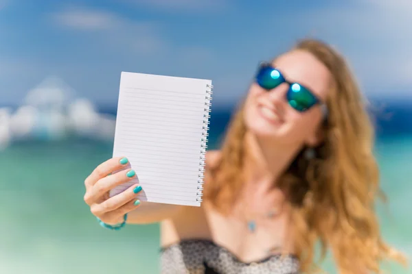 Giovane donna europea sorridente tiene in mano blocco note con mare tropicale turchese e barca sullo sfondo — Foto Stock