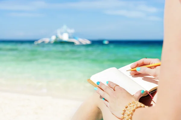 Europese vrouw zit op de kust van tropische turquoise zee en wrigting door pen in Kladblok op zonnige zomerdag — Stockfoto