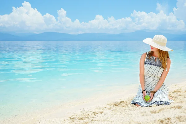 Jonge Europese vrouw in jurk, hoed en met groene appel zit op het zandstrand van rustige tropische zee op een zonnige dag — Stockfoto