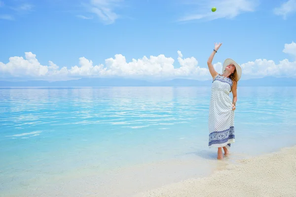 Unga europian kvinna i ljus klänning och hatt går på vit sandstrand nära lugna havet leker med grönt äpple på solig dag — Stockfoto