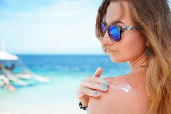 Mujer joven con gafas de sol applyng crema protector solar en su mano en la playa cerca del mar tropical turquesa bajo el cielo azul — Foto de Stock