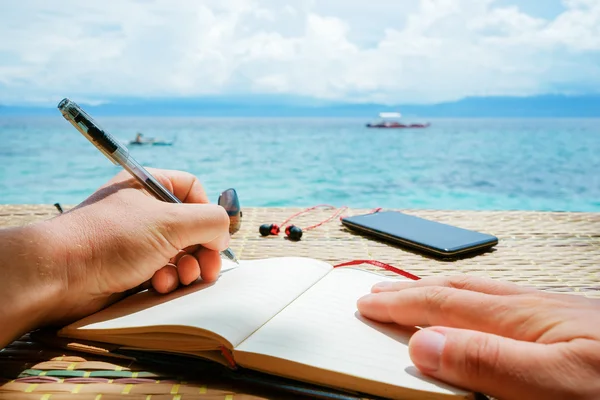 caucasian man is writing sime idea, message or letter in his notepad by pen while he sitting on the beach of tropical sea with boat