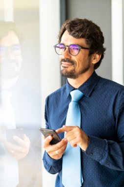 Gözlüklü bir iş adamı gülümsüyor, akıllı telefonunu tutuyor ve pencereden bakıyor. Mutlu başarılı erkek portresi 