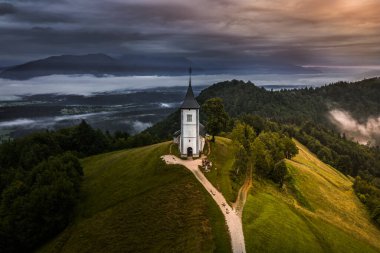Jamnik, Slovenya - Jamnik St. Primoz kilisesinde sisli bir yaz gündoğumunun insansız hava aracı görüntüsü. Sis, rengarenk gökyüzü ve arka planda Julian Alps olan küçük şapelin arkasına gider. 