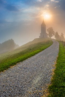 Jamnik, Slovenya - Jamnik St. Primoz kilisesinde küçük çakıl yolu ve yeşil çimenleri olan büyülü sisli bir yaz sabahı. Sis, mavi gökyüzü ve arka planda yükselen güneşle küçük bir şapelin etrafında dönüyor.
