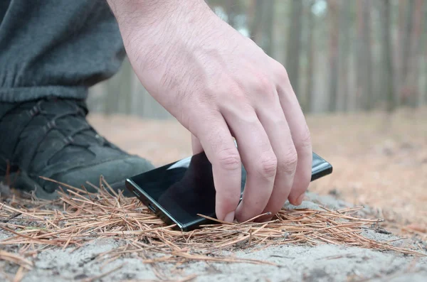 Male hand picking up a lost mobile phone from a ground in autumn fir wood path. The concept of finding a valuable thing and good luck