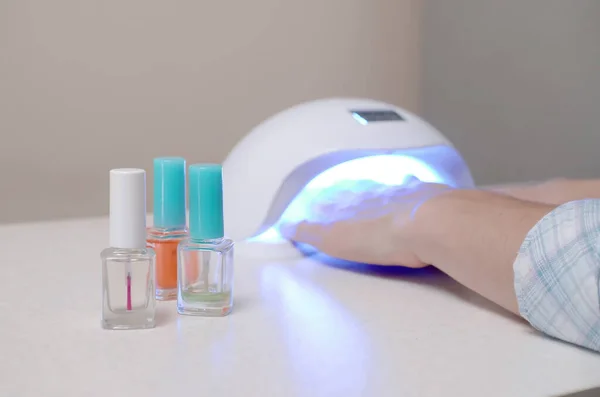 Three colorless transparent nail polish bottles in background of female hands in gel uv led nail white lamp for drying manicure. Beauty care women procedures in salon or home on gray background