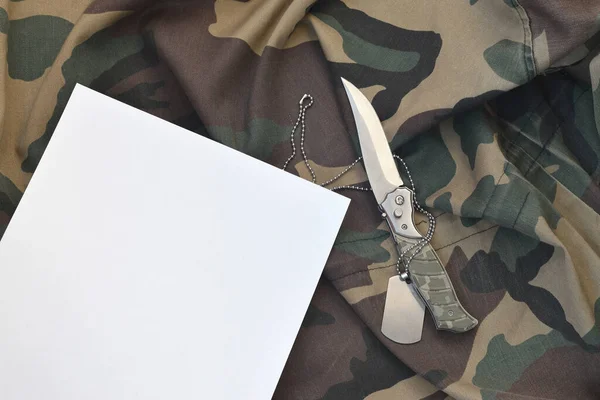 Blank paper with knife and army dog tag lies on camouflage military uniform. Copy space for notes during military service period