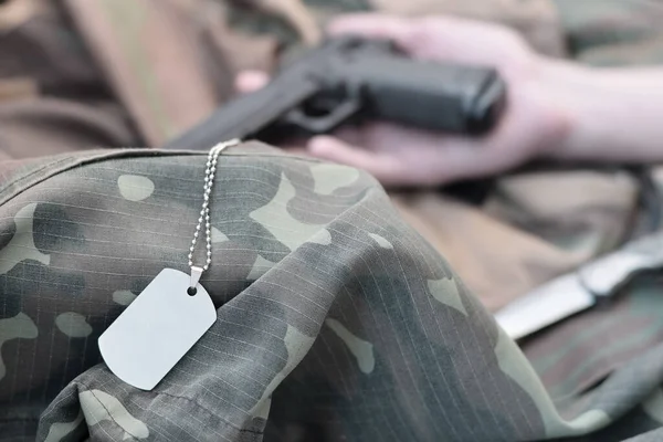 Dog tag with armed hand of dead recruit of Russian army lies on many camouflage clothes. Killed in action or suicide concept