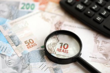 Brezilya reais banknotları ve büyüteçli hesap makinesi. Mevcut Brezilya gerçek para faturaları ve loupe ile calc büyük miktarda masada yatıyor. Brezilya 'da vergilendirme ve para konsepti