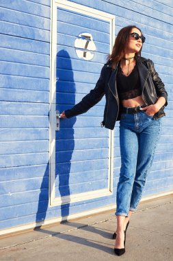 Açık poz güneş gözlüğü takmış uzun kıvırcık saçlı manken. Kot pantolon, deri ceket.
