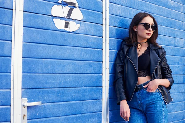 Model mit langen lockigen Haaren und Sonnenbrille, die im Freien posiert. Jeans, Lederjacke. — Stockfoto