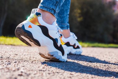 Asfalt bir yolda kot pantolon giymiş kadın bacaklarına siyah ayakkabılar takılmış beyaz spor ayakkabılar.