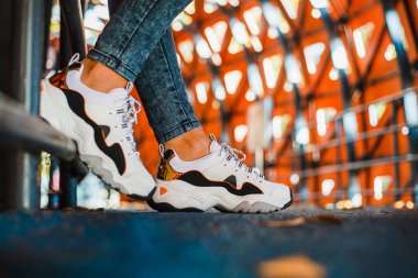 Kotlu, beyaz spor ayakkabılı, siyah pantolonlu demir çitin yanında duran bir kızın bacakları.