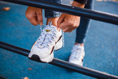 Beyaz spor ayakkabılarına bağcıklar bağlayan kadın elleri, siyah ayakkabılar, demir bir korkuluğun üzerinde duruyor.