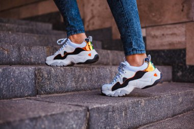 Sokakta mermer bir merdivende kot pantolon giymiş siyah bacaklı beyaz spor ayakkabılar.