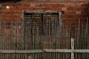 Dikey bambu çubuklardan yapılmış, kırmızı tuğlalı duvarın önünde duran ve el yapımı inşaat malzemelerini gösteren kırsal bir çit..