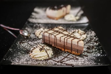 Portion of cake with three chocolates on a plate and a black background sprinkled with icing sugar, with cream and chocolate syrup