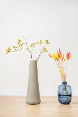 Uzatılmış gri bir vazoda yeşil yaban mersini dalları ve mavi cam sürahide renkli pankartlar.