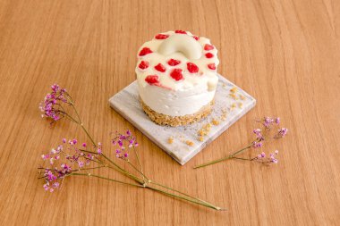 Üzerinde beyaz çikolata ve ahududu olan mini cheesecake, bisküvi toprağı, çiçekler ve küçük gri bir mermer tabağa yerleştirilmiş.