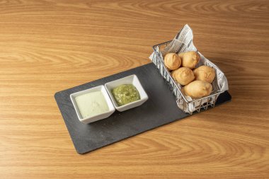 Small metal basket full of cheese sticks with two green sauce to dip on a black slate plate