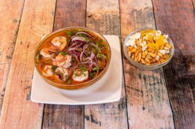 Bir cam kasede büyük miktarda karides ceviche ve yanında ahşap bir masa üzerinde Peru kankasıyla dolu daha küçük bir tane.