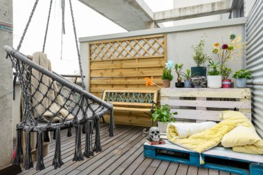 Teras köşesinde çeşitli bitkiler, boyanmış palet koltuklar, sarı şilteler ve ortasında kafatası var.