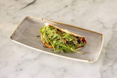 Buğday tortilla, domates, susam tohumu, roka ve gri tabakta küçük solucanlara benzeyen pilav.