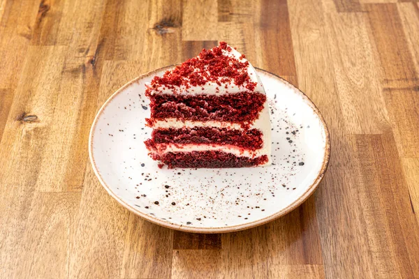 Red velvet cake portion with three layers of red sponge cake with white filling on white plate and wooden table