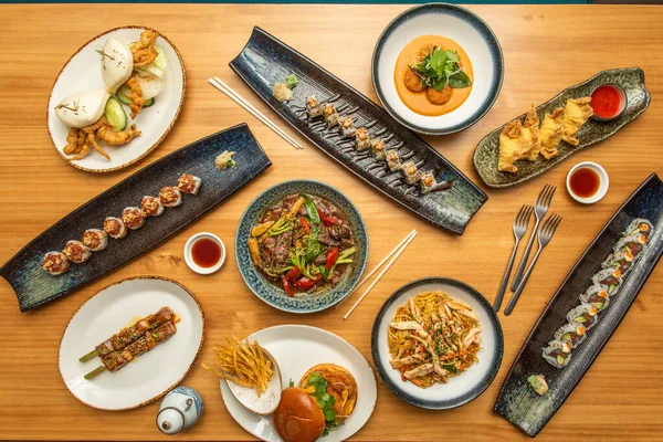 Set of beautiful asian food dishes on wooden table. Uramaki, tempura crab dip, fried gyozas, bluefin tuna skewer, stir-fry noodles, beef stew with vegetables, forks, chopsticks and soy sauce