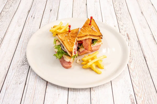 club sandwich with chicken, bacon, iceberg lettuce and ham, french fries, red onion, round white plate on white table.