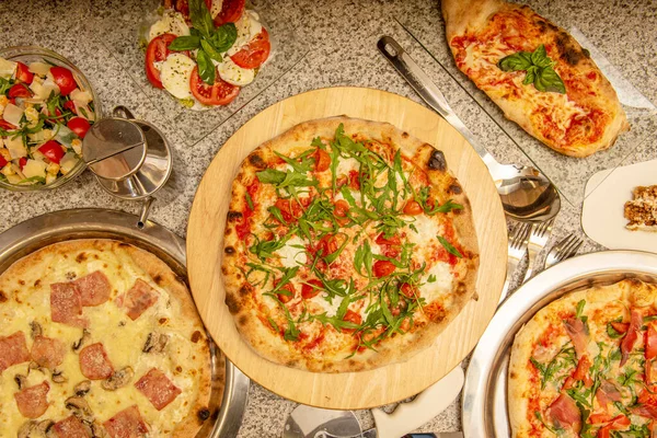 Pizzas and typical Italian dishes on a granite table. Margherita pizza with arugula, calzone with cheese, carbonara pizza with bacon, caprese salad