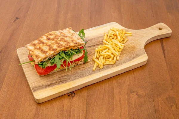 Focaccia sandwich with tomato, sliced buffalo cheese, dehydrated tomato and arugula in quantity with a garnish of fried potatoes on wooden board