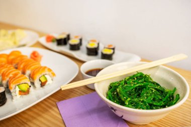 Bir kase Wakame salatası, ahşap yemek çubukları, makis ve uramaki arka planda bir Asya restoranının masasında.
