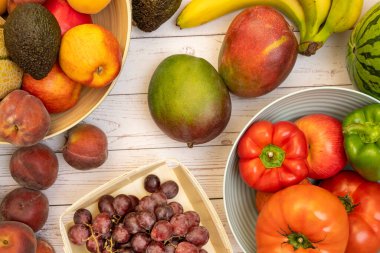 Mevsimlik meyve ve sebzeler beyaz masa ve kaselerde. Fildişi rengi mango, kırmızı biber ve domates, olgun muz, kırmızı elma ve şeftali, yeşil avokado ve karpuz.