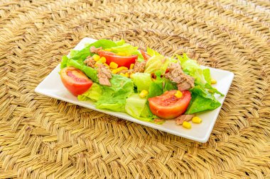 Akdeniz salatası, büyük domates parçaları, tatlı mısır, buzdağı marulu ve beyaz tabakta konserve kuzey bonitosu.