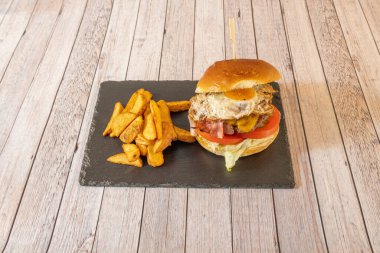 Classic beef burger with fried egg, sliced fried bacon, melted cheddar cheese, tomato slices and pickles and lettuce leaves garnished with fried potato wedges