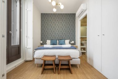 Büyük boy yatak, tahta tabureler, mavi yastıklar, beyaz dolaplar ve arka planda kiralık apartman dairesi.