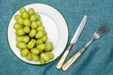 Yeşil kenarlı, mavili metal tabakta beyaz tatlı üzümleri ve mavi kumaş üzerinde klasik çatal bıçak takımı.