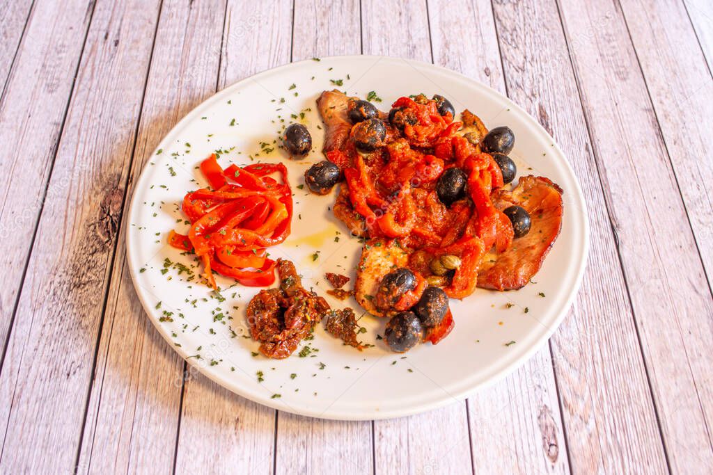 Pork escalopes recipe with dehydrated tomatoes, piquillo peppers, black olives and oregano on a white plate