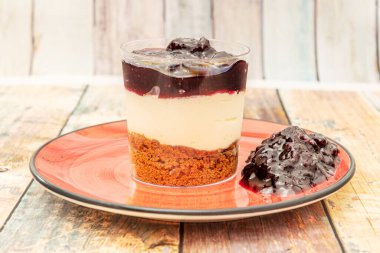 Kristal bardaktaki tatlı, kırmızı tabakta bisküvi, cheesecake ve yaban mersini reçelini gösteriyor.