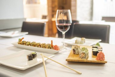 Sushi uramaki dürümü, ginseng, wasabi, yemek çubukları ve beyaz masa örtüsü olan bir restoranda bir bardak şarap.