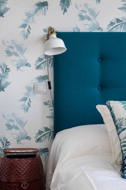 Kiralık bir dairenin yatak odasında beyaz lamba ve beyaz çarşaflı mavi kumaş yatak başlığı.