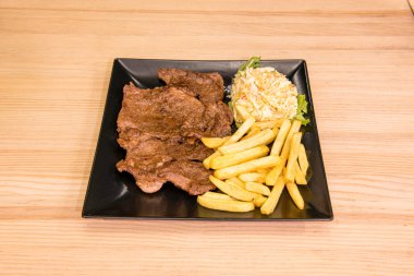 Biftek, patates kızartması ve lahana salatasıyla süslenmiş siyah kare tabakta.